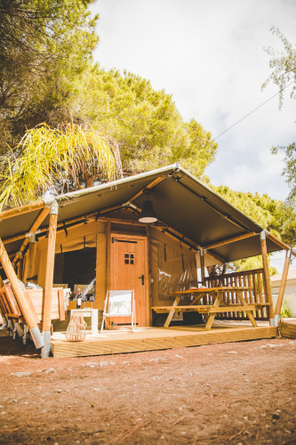 Das Safari-Zelt Glamping Safari Mistral mit Holzveranda und Picknickbank in einem waldreichen Gebiet.