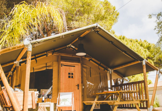 Glamping Safari Mistral safari tent with wooden deck, picnic table, and chair, set among lush green trees.