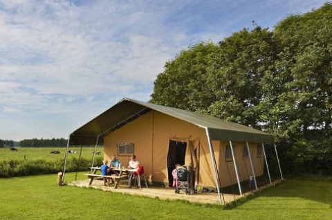 Gezin zit aan picknicktafel voor een safaritent op een groene weide met bomen in de achtergrond.