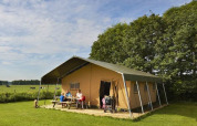 Familie sidder ved picnicbord foran en stor safaritelt på en grøn mark med træer i baggrunden.