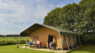 Familie sidder ved picnicbord foran en stor safaritelt på en grøn mark med træer i baggrunden.