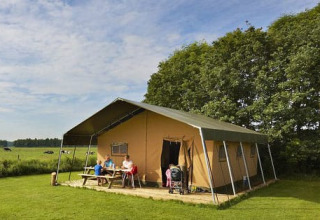 Family sits at a picnic table outside a large safari tent on a grassy field with trees in the background.