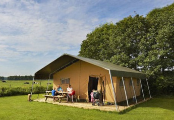 Gezin zit aan picknicktafel voor een safaritent op een groene weide met bomen in de achtergrond.