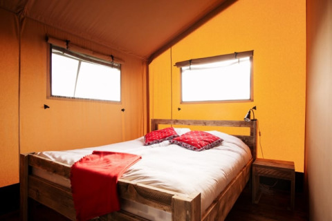 Chambre confortable sous tente safari avec lit en bois, draps blancs, coussins rouges et lumière naturelle.