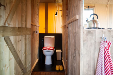 Binnenzicht van een safaritent met toilet met rood deksel, houten wanden en fluitketel in de keuken.