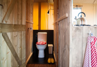 Vue intérieure d’une tente safari avec toilettes à siège rouge, murs en bois et bouilloire dans la cuisine.