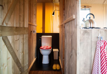 Interior view of a safari tent with a toilet featuring a red seat, wooden walls, and a tea kettle in the kitchen.