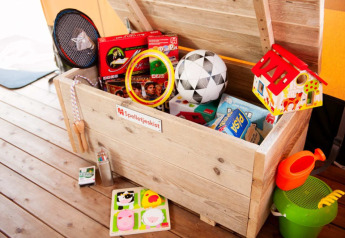 Hölzerne Spielzeugkiste in einem Safarizelt mit Brettspielen, Sportgeräten, einem Spielzeughaus und Strandspielzeug.