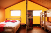 Interior de tienda safari con cama doble, literas de madera y mobiliario rústico bajo lona amarilla.