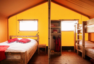 Interior of a safari tent featuring a double bed, bunk beds, and wooden furniture beneath a yellow canopy.