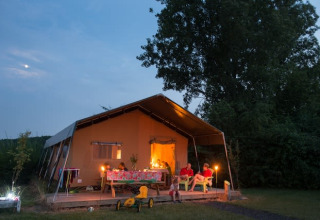 A family enjoying a cozy evening in front of a safari tent, glowing warmly under the trees at dusk.
