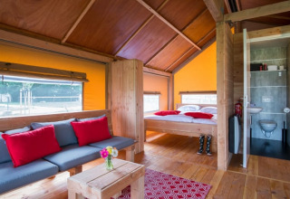 Interior de la tienda safari Haytent con muebles de madera, sofá, cama, baño y cojines rojos.