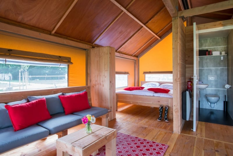 Interior de la tienda safari Haytent con muebles de madera, sofá, cama, baño y cojines rojos.