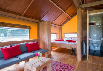 Interno della tenda safari Haytent con mobili in legno, divano, letto, bagno e cuscini rossi.