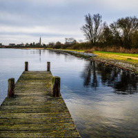 Muelle de madera junto a lago tranquilo, árboles y zona de glamping en parque vacacional en día nublado.