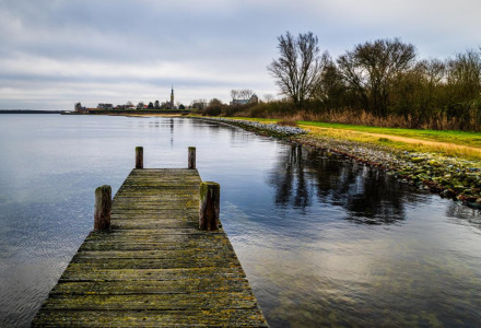 Muelle de madera junto a lago tranquilo, árboles y zona de glamping en parque vacacional en día nublado.