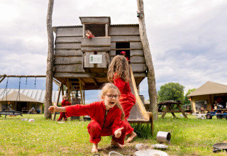 Kinderen spelen in rode pakken bij een houten huisje op FarmCamps Op de Beemd in Noord-Brabant, Nederland.