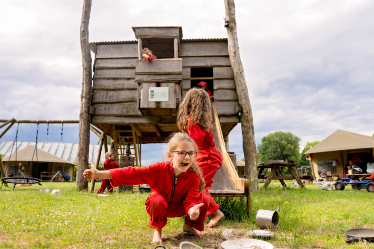 Kinder spielen in roten Overalls vor einem Holzhaus bei FarmCamps Op de Beemd in Nordbrabant, Niederlande.