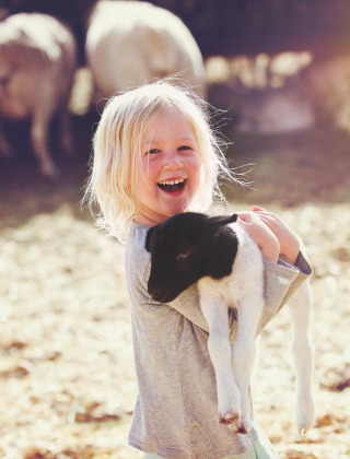 Niña rubia sonriente sostiene un cordero blanco y negro, con ovejas pastando al fondo en un día soleado.