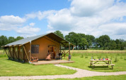 Safari telt ved navn Lodgetent på en grøn mark med piknikbord, træer i baggrunden og blå himmel.