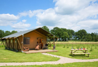 Safari-Zelt namens Lodgetent auf einer Wiese mit Picknicktisch, Bäumen im Hintergrund und blauem Himmel.
