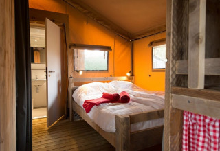 Interieur van een knusse safaritent met een houten bed, fris wit beddengoed, rode handdoeken en zicht op een kleine badkamer.