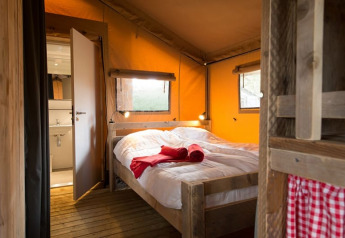 Interieur van een gezellige safaritent met een houten bed, wit beddengoed, rode handdoeken en uitzicht op een kleine badkamer.