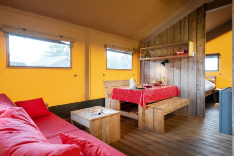 Interior of a safari tent with wooden furniture, red tablecloth, cozy sofa, and warm yellow walls.