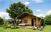 Safaritent op een groene weide met terras, tafel, stoelen en kinderspeelgoed onder een zonnige lucht.