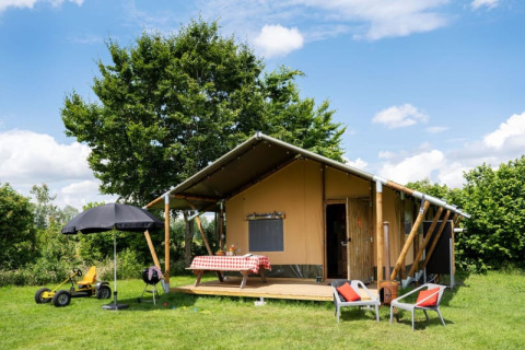 Safari-Zelt auf einer Wiese mit Terrasse, Tisch, Stühlen und Kinderspielzeug unter blauem Himmel.