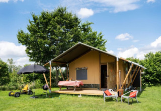 Tente safari sur un champ verdoyant, terrasse, table, chaises et jouets d’enfants sous ciel ensoleillé.