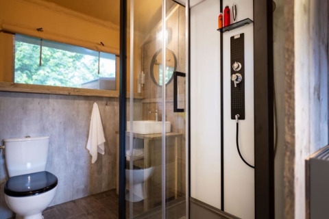 Salle de bain dans une tente safari avec douche moderne, toilettes, lavabo et grande fenêtre lumineuse.
