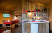 Interior de tienda safari con cocina de madera, mesa de comedor y detalles en rojo bajo luz cálida.