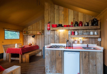 Binnenkant van een safaritent met houten inrichting, compacte keuken en eetruimte met rood tafelkleed.