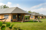 Safaritenten op een grasrijke camping met hangmatten, picknicktafels en terrasstoelen onder een blauwe lucht.
