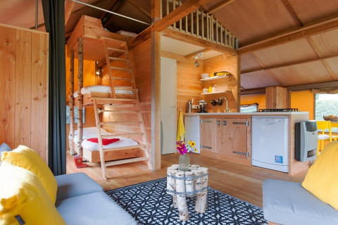 Interno di una tenda safari con arredi in legno, cucina, letti a soppalco e zona lounge accogliente.