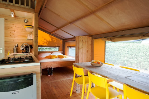 Vista interior de una tienda safari con cocina, mesa de comedor de madera, sillas amarillas y cama doble.