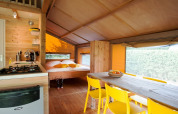 Vista interior de una tienda safari con cocina, mesa de comedor de madera, sillas amarillas y cama doble.