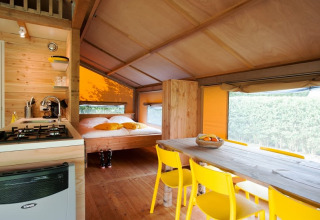 Vista interior de una tienda safari con cocina, mesa de comedor de madera, sillas amarillas y cama doble.