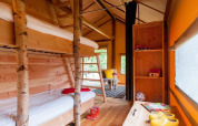 Intérieur de tente safari avec lits superposés, décor en bois, coin salon confortable et lumière naturelle.