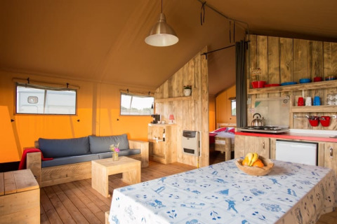 Binnenzicht van een safaritent met houten meubels, een sofa, eettafel en kleine keukenhoek.