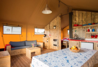 Binnenzicht van een safaritent met houten meubels, een sofa, eettafel en kleine keukenhoek.