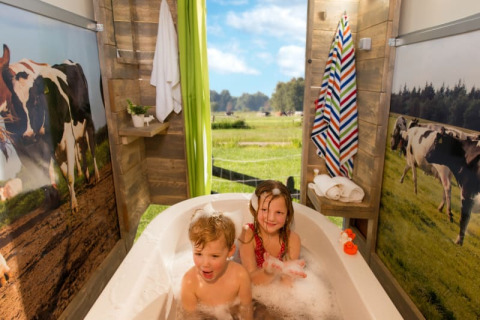 Twee kinderen genieten van een bad in een safaritent met koeiendecor en uitzicht op een weide met koeien.