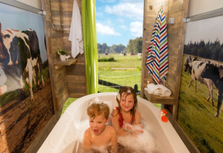 Twee kinderen genieten van een bad in een safaritent met koeiendecor en uitzicht op een weide met koeien.