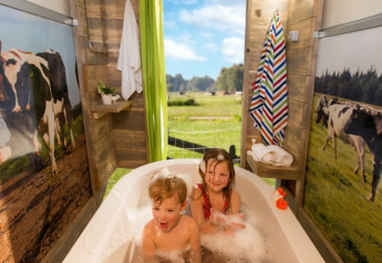 Twee kinderen genieten van een bad in een safaritent met koeiendecor en uitzicht op een weide met koeien.