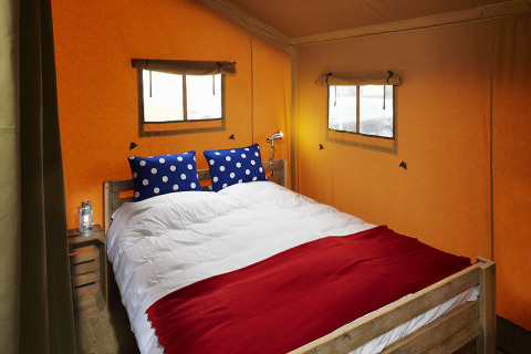 Interior de tienda safari con cama doble, cojines azules de lunares y aire acondicionado.