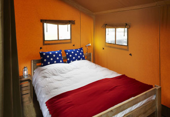 Interior de tienda safari con cama doble, cojines azules de lunares y aire acondicionado.