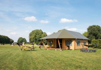 Safaritent op een groene weide met picknicktafel, hangmat, vuurplaats en koeien onder een blauwe hemel.