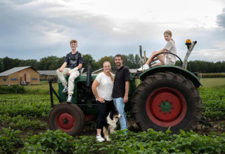 Famille et chien posent près d’un tracteur sur une ferme verte à Limburg, Pays-Bas, avec cabanes au fond.