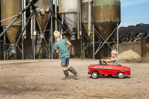 Dos niños juegan en una granja, uno tira del otro en un coche de juguete rojo frente a unos silos grandes.